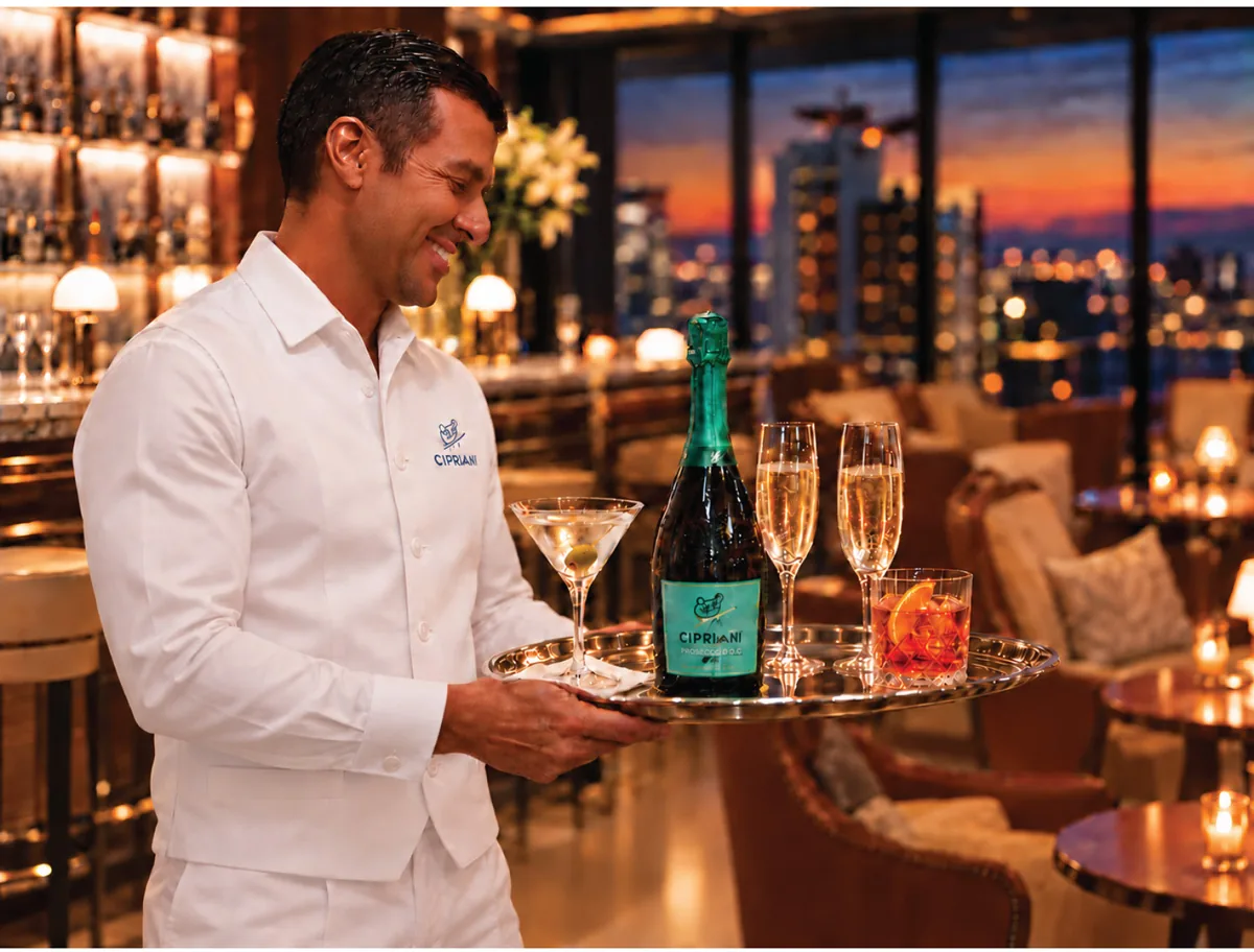 Imagen ilustrativa de un mesero con chaqueta blanca que lleva una bandeja de plata con cócteles, copas de champán y una botella de prosecco por un salón al anochecer.