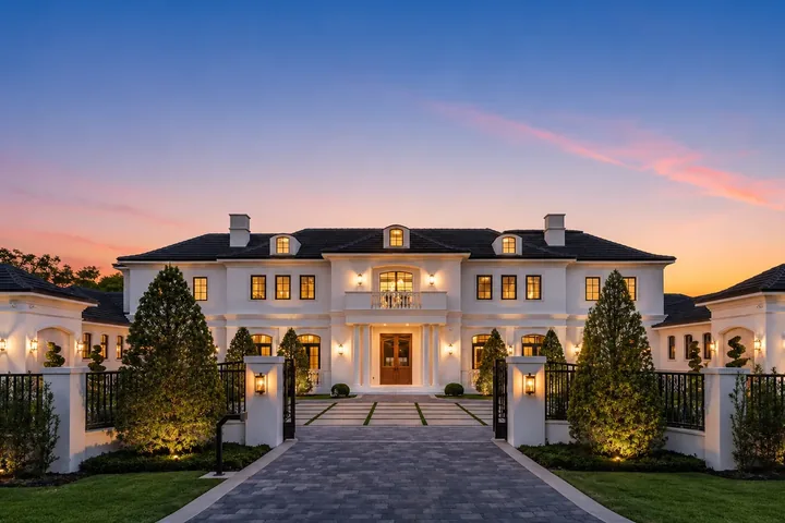 Residencia de lujo en Pinecrest al atardecer, con pórtico iluminado y entrada adoquinada con portón en un terreno amplio y ajardinado del Village of Pinecrest, Florida