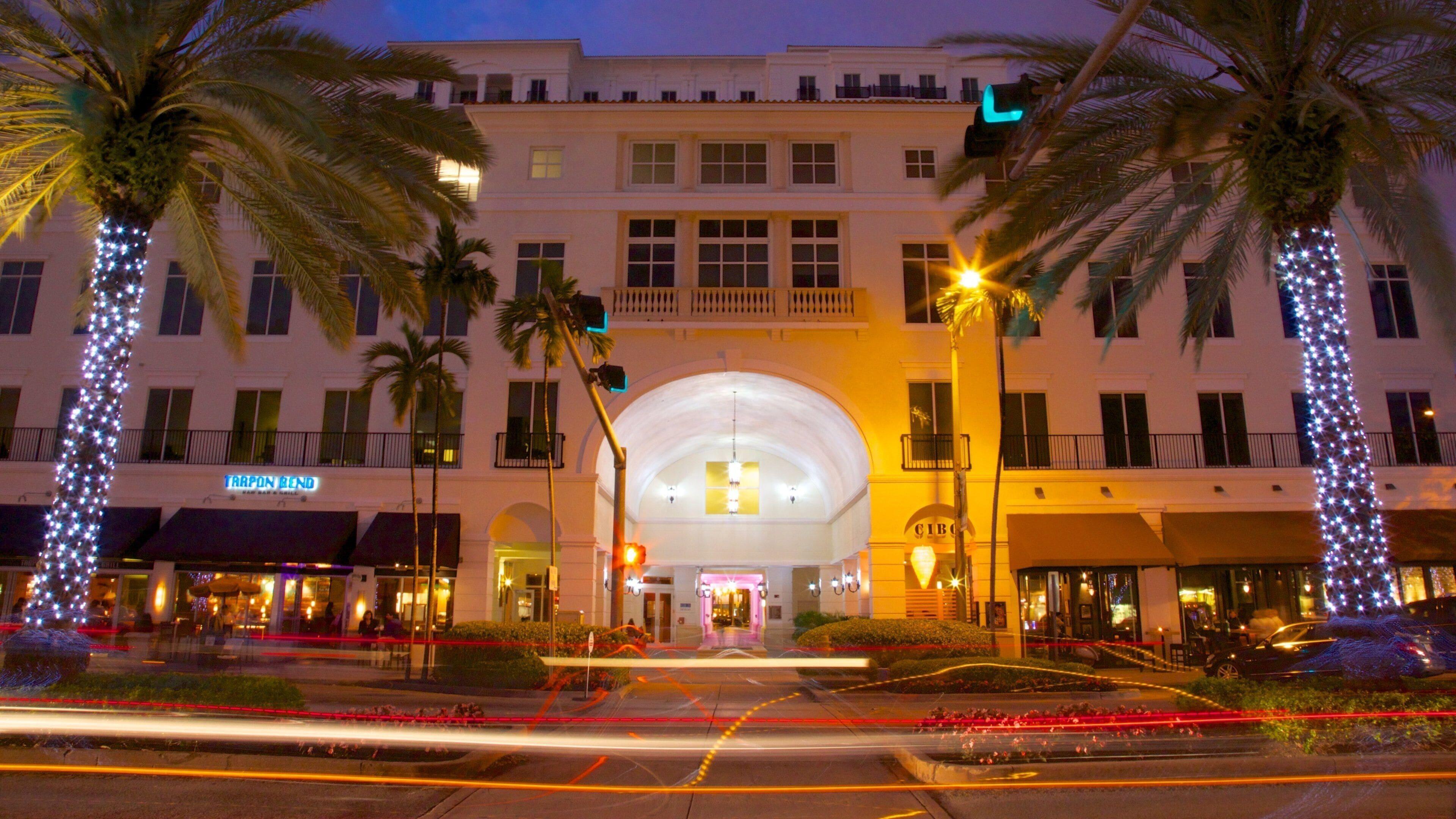 Una calle de Coral Gables de noche bordeada de palmeras y vitrinas iluminadas cerca de The Avenue Coral Gables