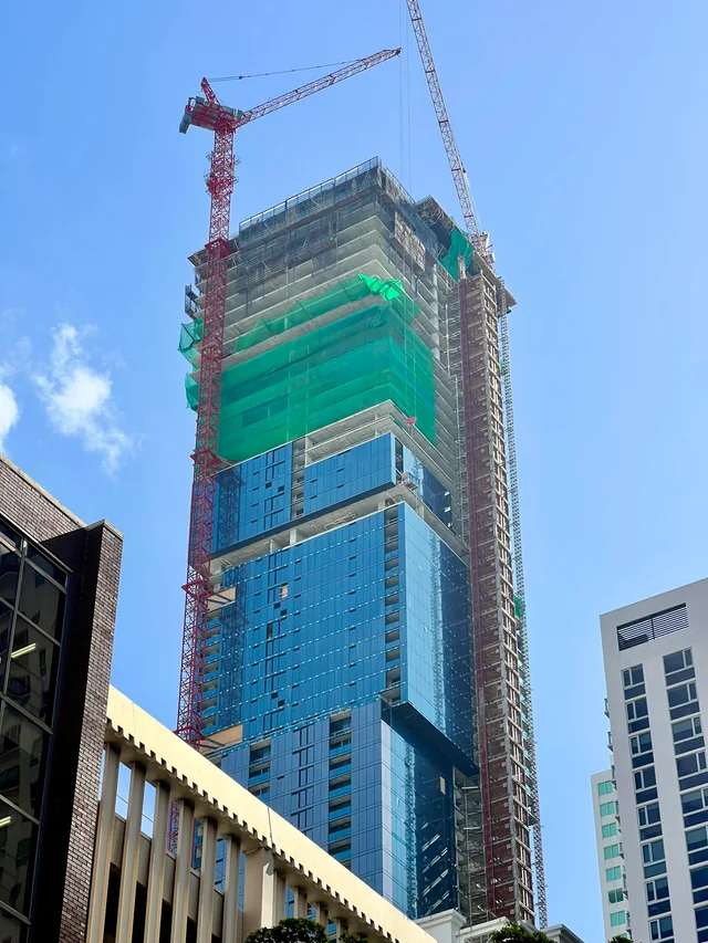 Close view looking up at Waldorf Astoria Miami in August 2026 showing installed dark glass curtain wall on the lower cubes and green protective netting around the active concrete work above